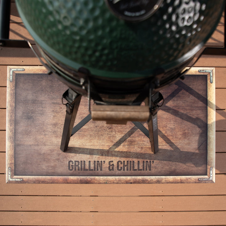 top-down view of the customized grill mat with a woodgrain pattern and text that reads ”Grillin’ & Chillin’” and a green grill on top of it 